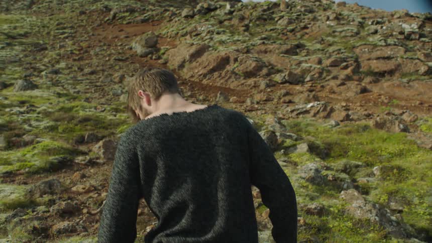 A man with a sweater walking up a rocky hill with the sky and clouds in the background