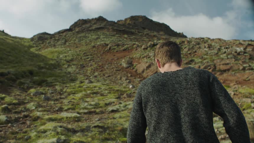A man with a beard and wearing a gray sweater walks up a hill during the day
