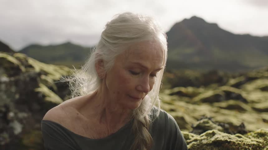An elderly caucasian woman sitting and touching some moss with mountains behind her