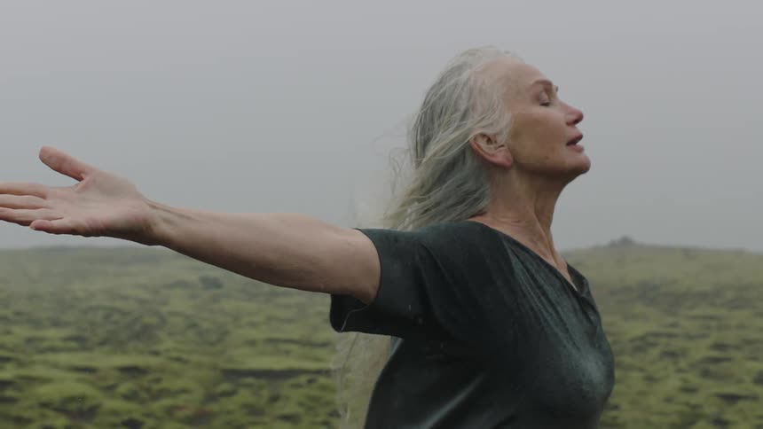 Elderly caucasian woman surrounded by grasslands outstretching her arms and feeling the wind on a cloudy day