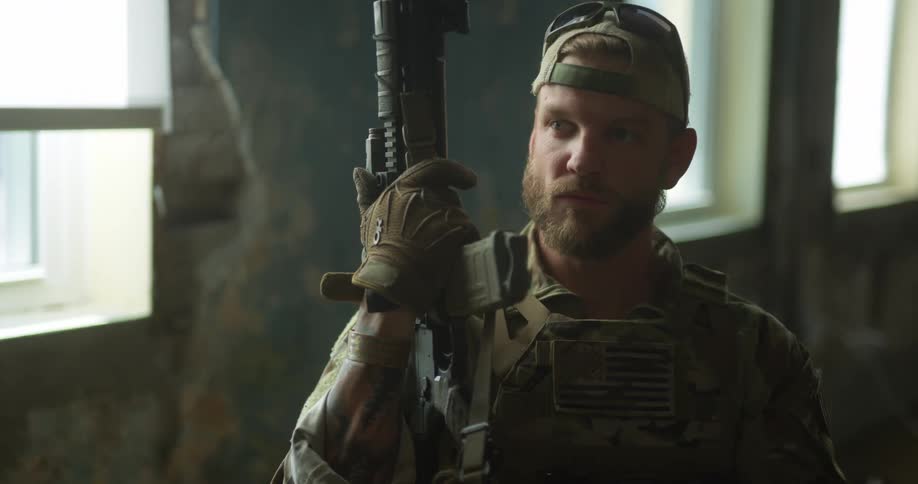 A soldier in combat uniform holds a firearm with one arm while sitting inside a ruined building during the day