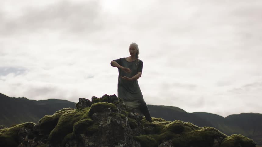 Woman stands on mossy rock in hilly area doing tai chi