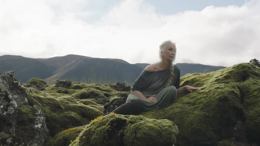 An elderly woman looks into the distance while resting atop a rock formation covered in moss