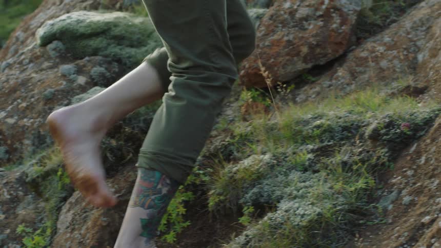 Young tattooed man climbs barefoot on the moss covered rocks of a hill