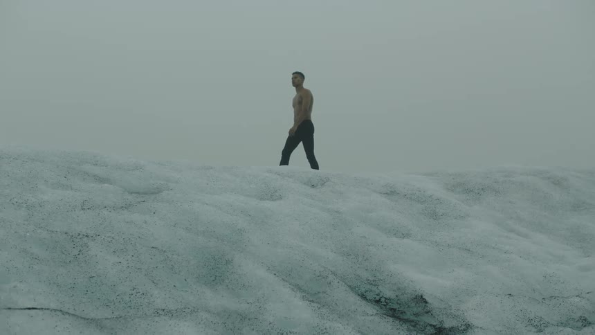 A shirtless young man walks along the top of a hill covered in snow under a cloudy sky