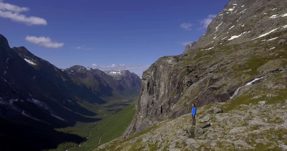 A person in a blue jacket stands on a rock on a mountain edge and overlooks a valley
