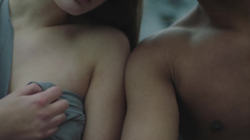A naked young man and a young woman wrapped in a blanket sit side by side with their heads together outdoors while staring into the camera
