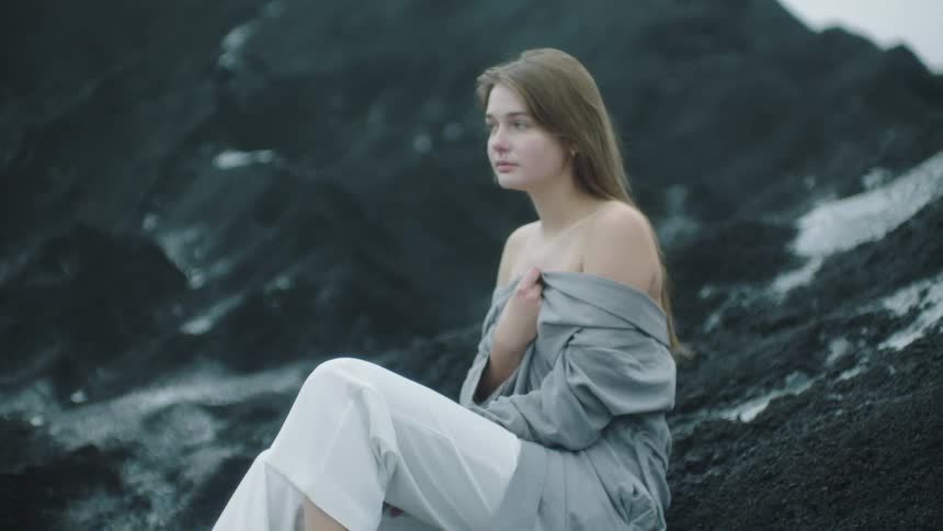 Young blonde woman wearing a white dress and gray robe sitting on the black sand