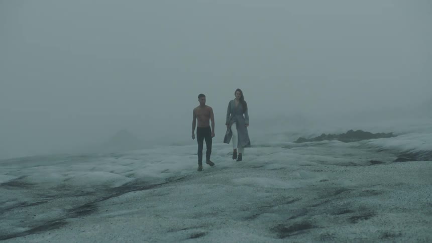 A man without a shirt on walks next to a woman across a snow covered field