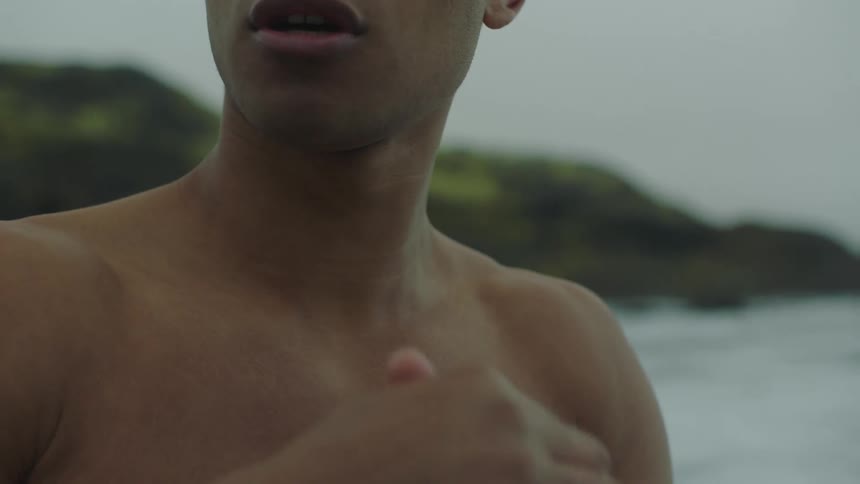 A shirtless young man stands on a shoreline with ice below him and rubs his hands together in the cold