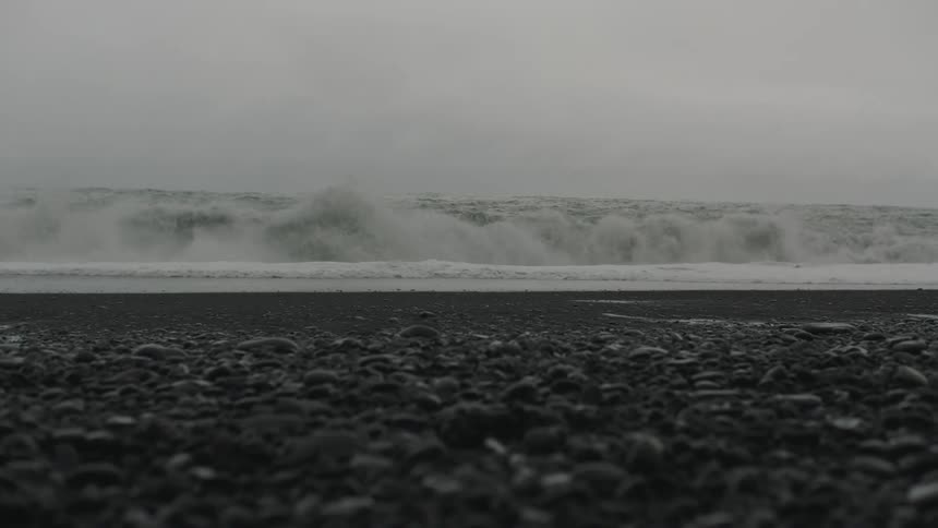 Ocean waves wash into a beach covered in a gravel under a cloudy gray sky