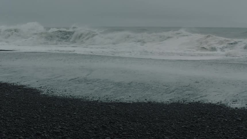 Large waves crash upon a beach that is lined with many small stones