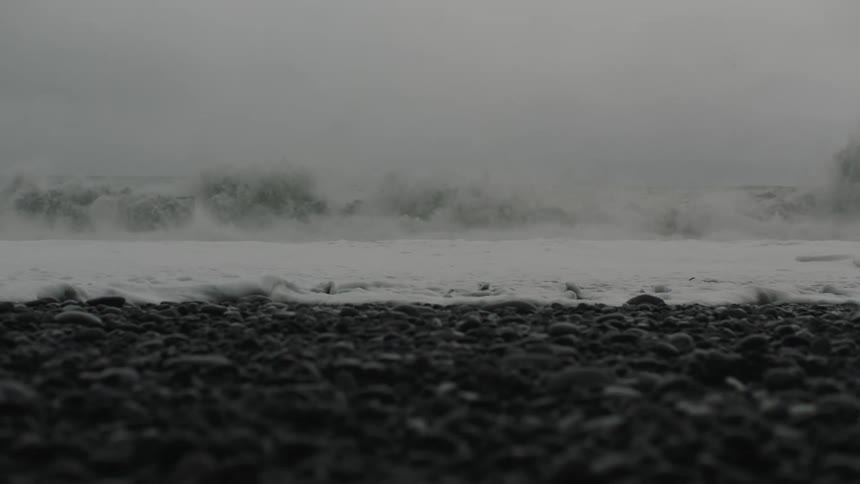 A beach with black sand and stones with the waves washing up on the shore with the sky and clouds in the background