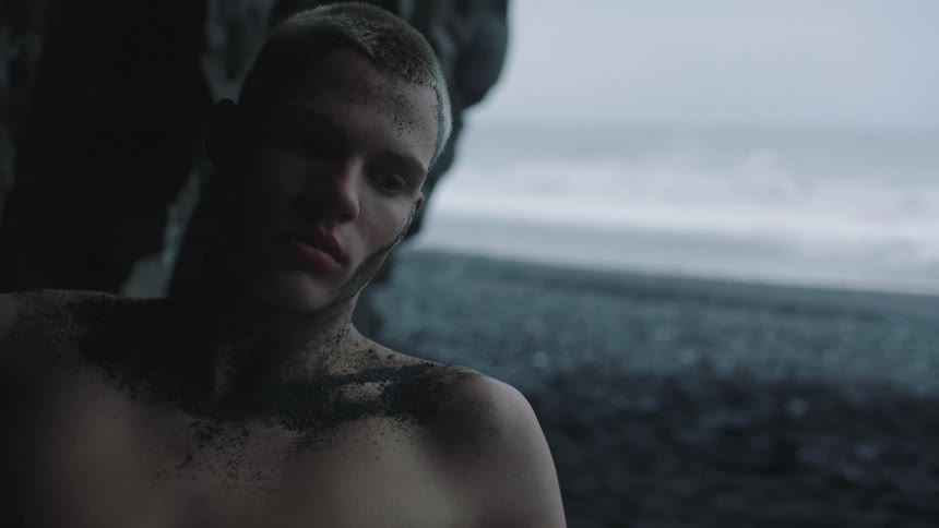 Young man looking at the camera reclined on a dark rock covered in black sand next to a beach