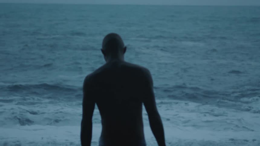 A shirtless young man in swim trunks slowly walks along the beach towards the incoming ocean waves