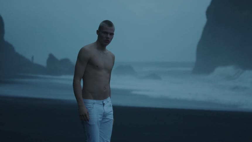 A bare chested man wearing jeans walks backwards on a beach in the evening