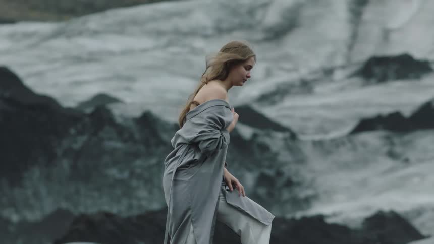 A woman with a gray robe walking with a cliff in the background
