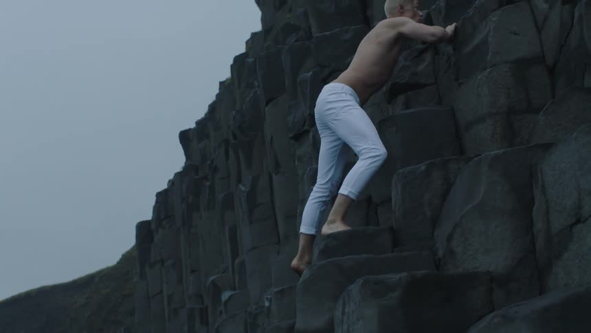 A man with white pants climbing a rocky cliff with the sky and clouds in the background