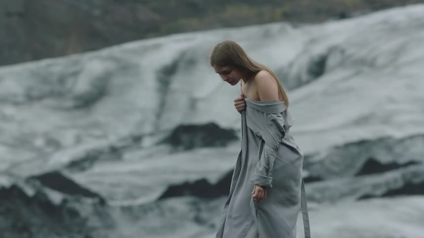 Young woman wearing a white dress and a grey coat walks over the rocks in the mountain