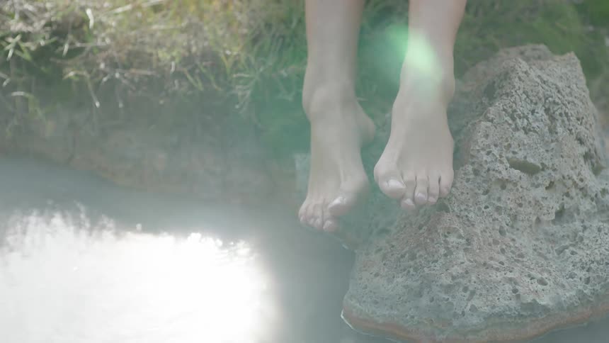 Steam flows over a pair of feet as a person rests them on a stone in a hot spring during the day