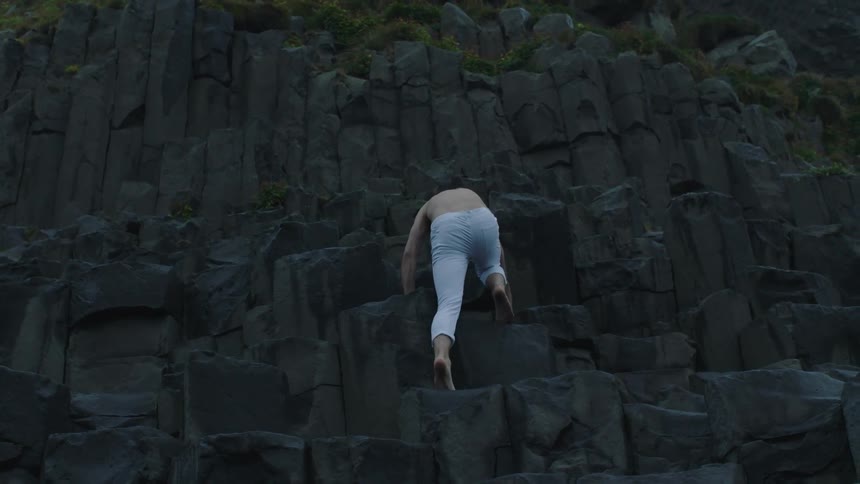 Young shirtless man climbs barefoot the black rocks on a steep cliff on the side of a mountain