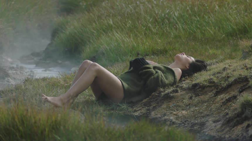 A woman in a large sweater rests in the grass amid steam from hot springs in a field during the day