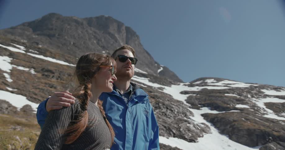 A man and woman stand at the base of a mountain looking forward and talking