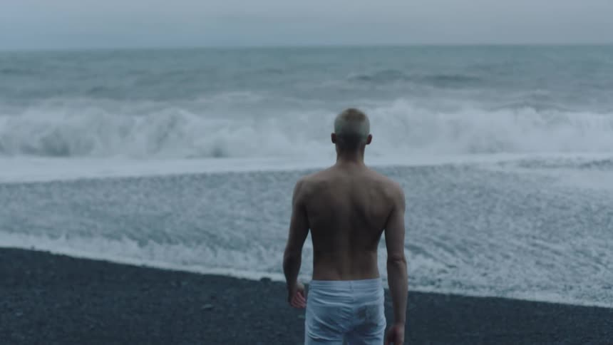 A shirtless man wearing white pants slowly walks towards the crashing waves of the ocean