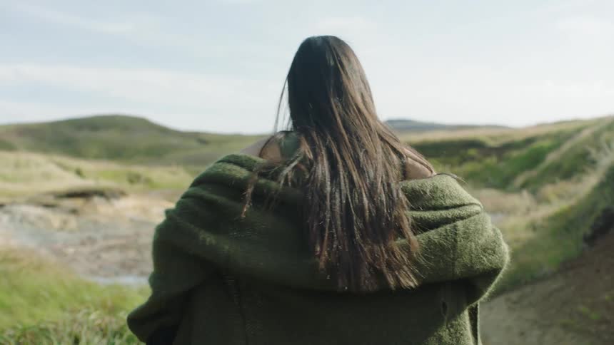 Woman wearing a thick green wool coat walking among the green small hills outdoors