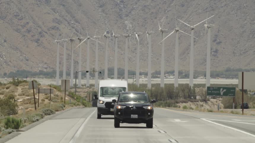 Traffic travels away from a wind farm on a road through an arid countryside during the day