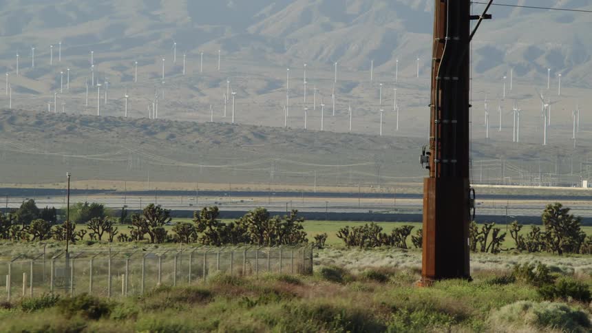 A grassy plain with a fence trees and a utility pole with windmills and hills in the background