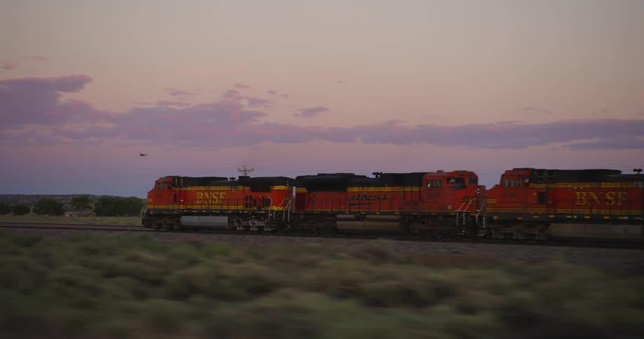 An orange and yellow train moves down tracks in a remote area of fields at dusk