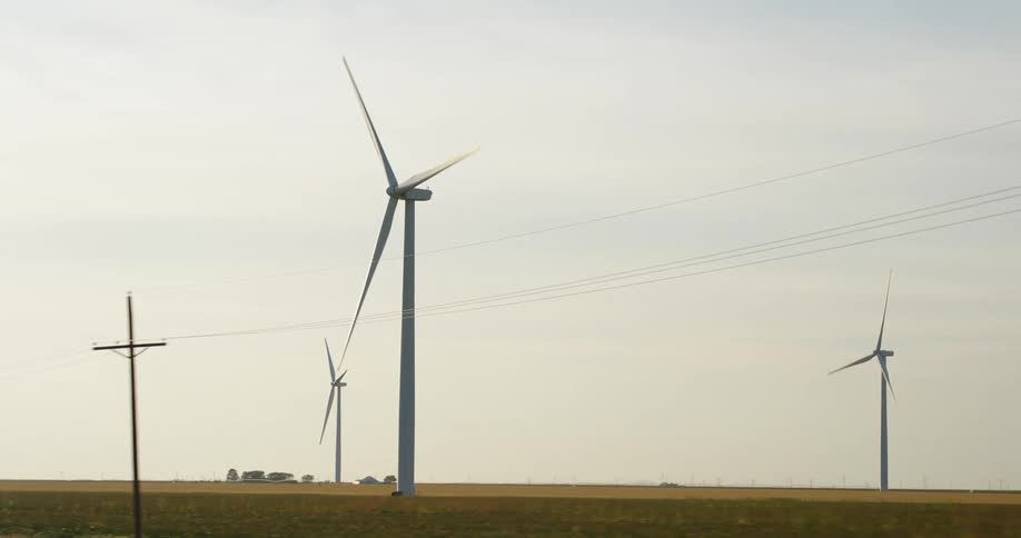 A wind farm is on a grassy field with overcast skies and a structure in the distance