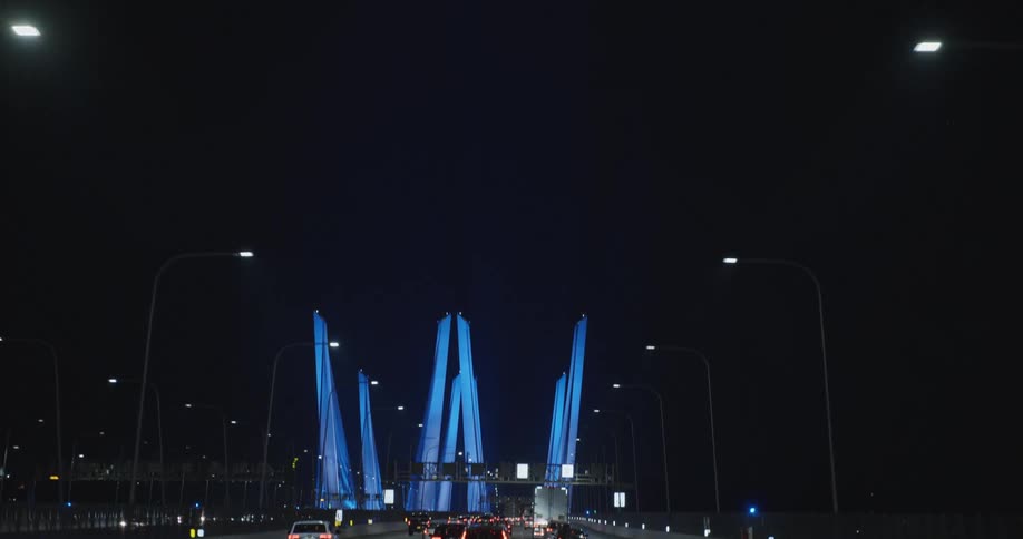 Colorful architecture looms in the distance over traffic traveling along a long highway bridge at night