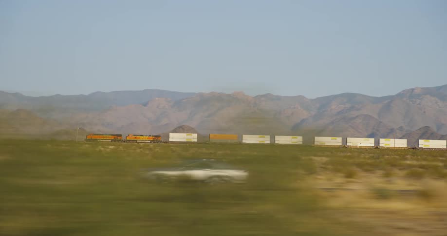 Trucks driving on a rural road passing in front of a freight train near the mountains