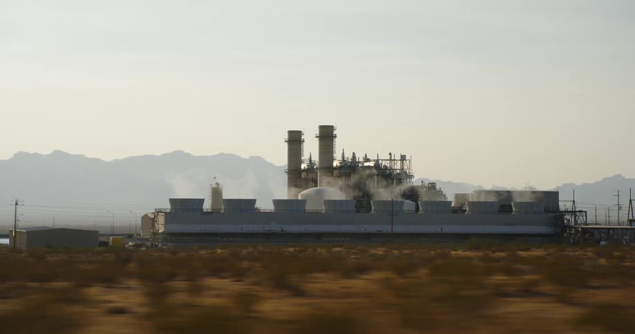 A factory with smoke with a grassy field in the foreground and a mountain range the sky and clouds in the background