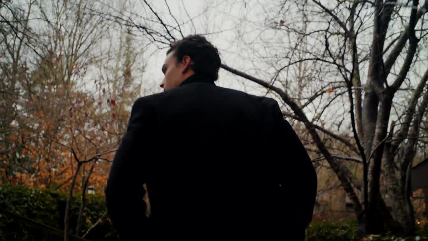 A man walks outdoors in a neighborhood with trees and foliage wearing a black jacket