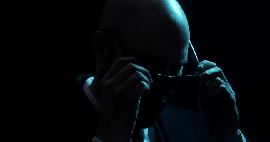 Bald man standing in a dark room puts sunglasses on his face