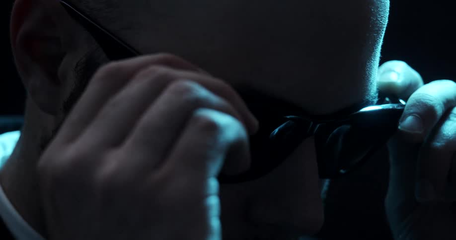 A man with facial hair places sunglasses on his face in shadows