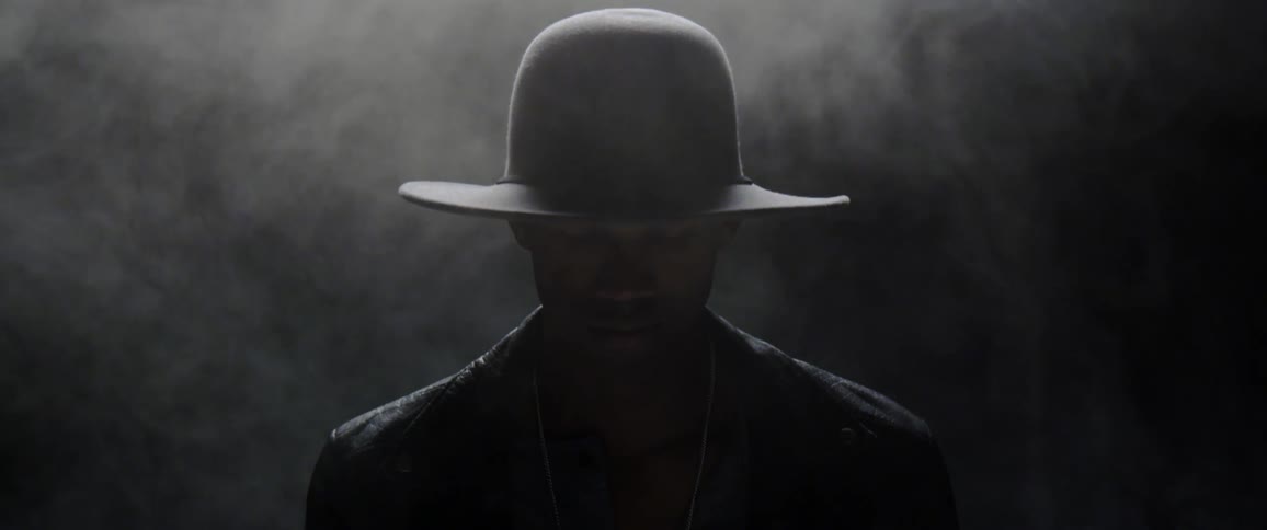 A man wearing a cowboy hat stands in the darkness as smoke drifts around him
