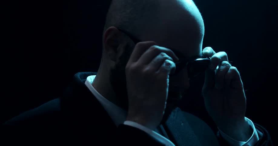 A bald man wearing a suit stands in the darkness and puts on glasses