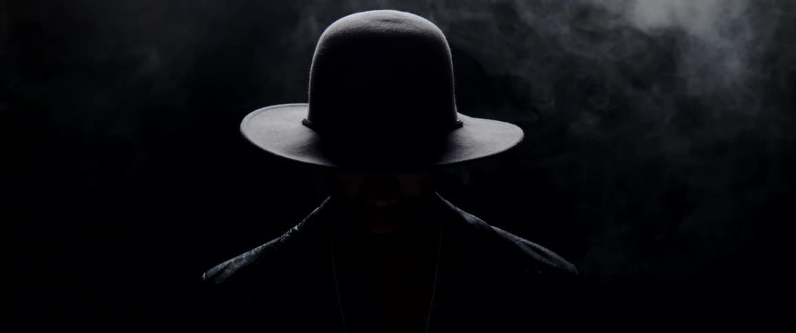 Man stands in dark with over head light smoking looks down and then up