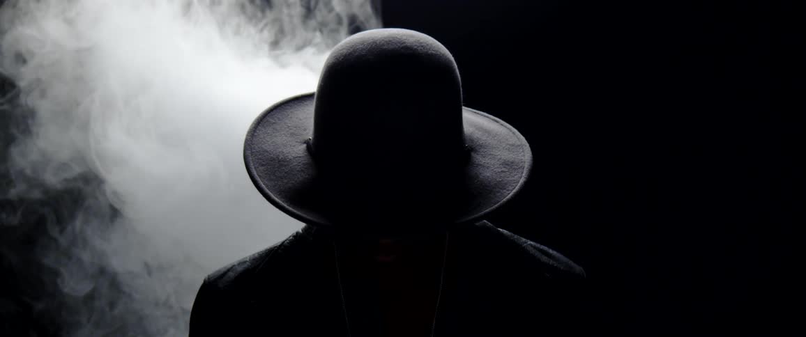 Silhouette of the back of a person wearing a wide brim hat in front of a cloud of white smoke in a dark room