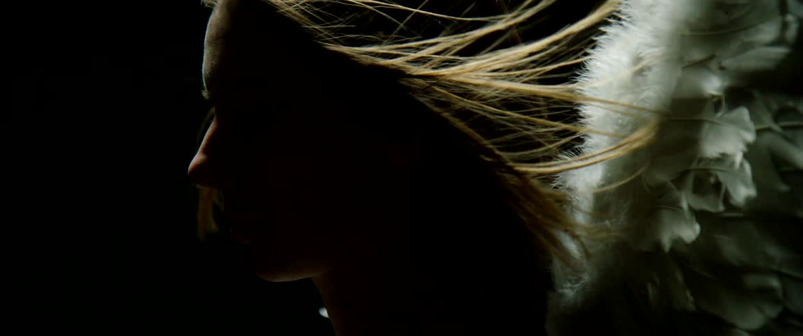 A woman stands outside with feathers attached to her back as her hair blows in the wind