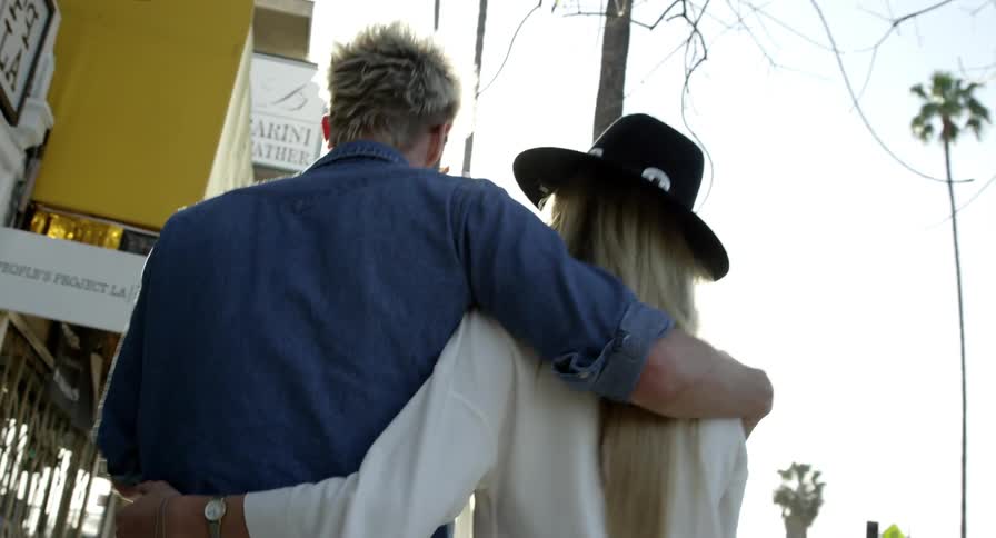 A man and a woman walking on the sidewalk hugging with trees the sky and clouds in the background