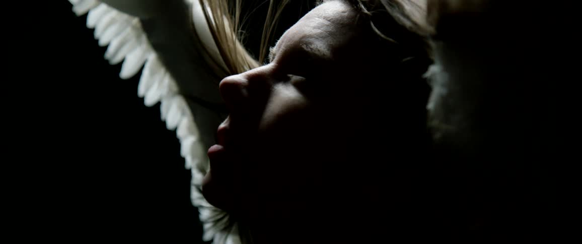 A female with angelic wings stands in the darkness looking upwards