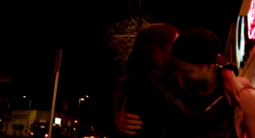 A woman kisses a man on the cheek as they embrace while walking down a sidewalk in a city at night