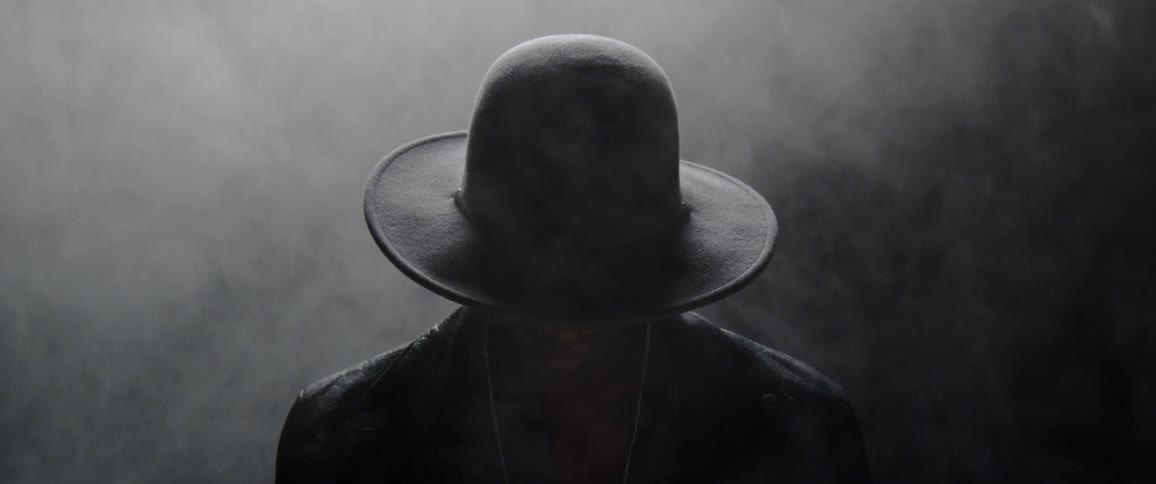 A mysterious person wearing a hard stands in a darkened location while surrounded by fog