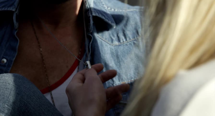 A woman sits with a man and touches the necklace around his neck while outdoors in the sun
