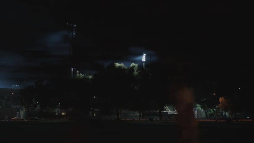 The surrounding exterior of a lighted stadium with trees and a parking lot at night